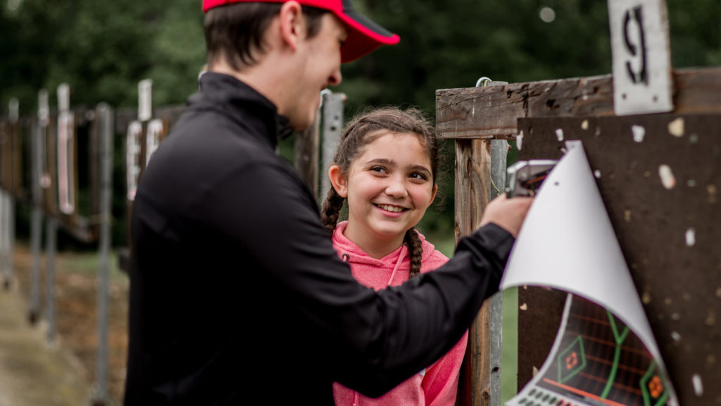A father showing his daughter how to staple a paper target to a target board.
